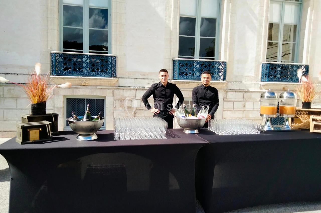 Deux serveurs en tenue noire derrière une table dressée avec boissons, verres et décorations, devant un bâtiment ancien.