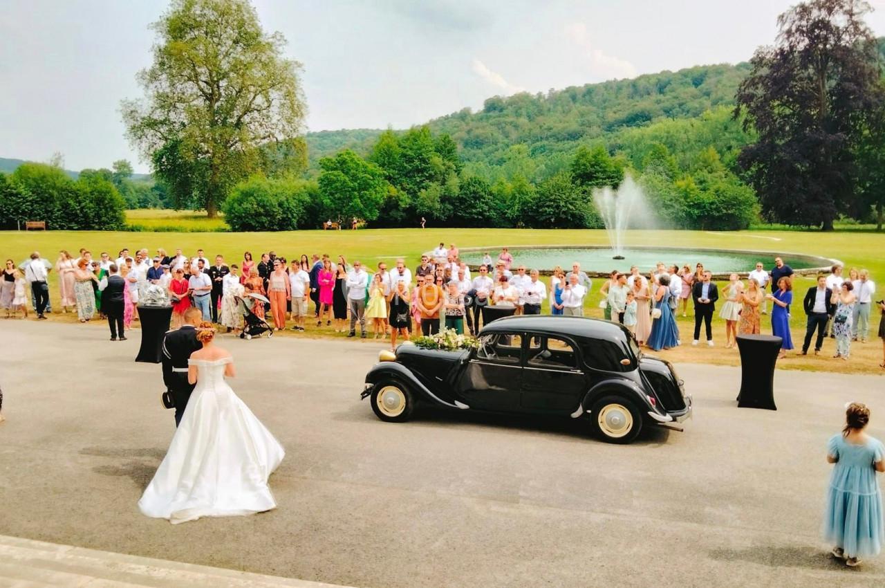 Un couple de mariés devant une voiture ancienne, entouré d'invités dans un parc avec une fontaine en arrière-plan.