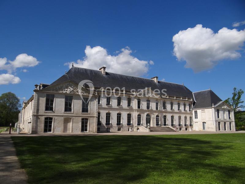 Façade d'un château en pierre avec toit en ardoise, entouré d'une pelouse et d'arbres sous un ciel bleu avec des nuages.
