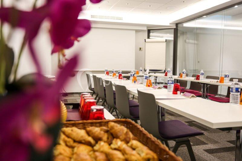 Salle de réunion préparée avec des tables disposées en U, bouteilles d'eau, blocs-notes et viennoiseries au premier plan.