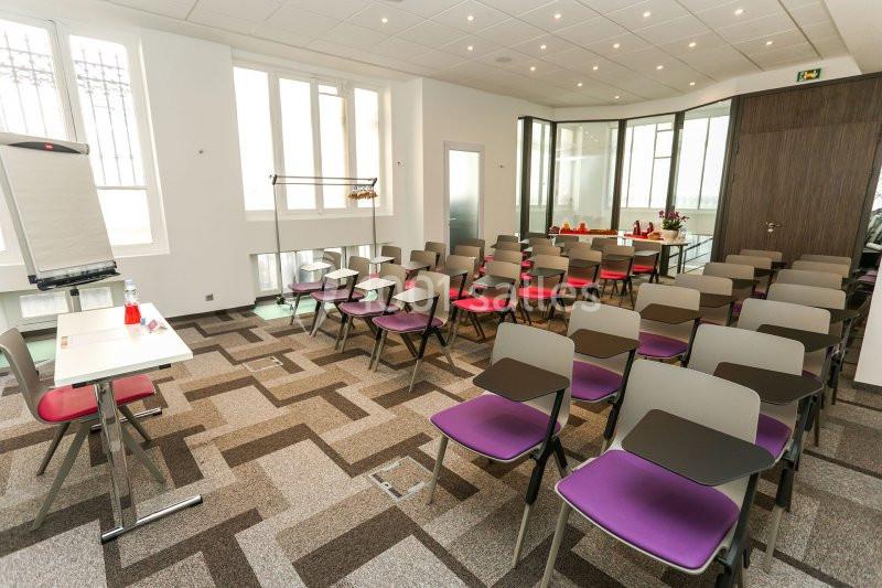 Salle de réunion lumineuse avec rangées de chaises colorées, un tableau de conférence et une table à l'avant.