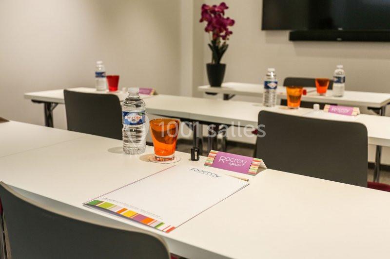 Salle de réunion avec tables alignées, blocs-notes, bouteilles d'eau, verres colorés et une orchidée en arrière-plan.