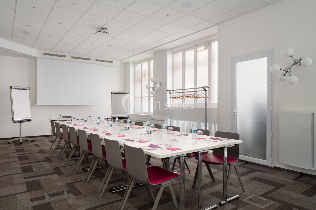 Salle de réunion lumineuse avec table rectangulaire, chaises, paperboards, écran de projection et porte-manteaux.