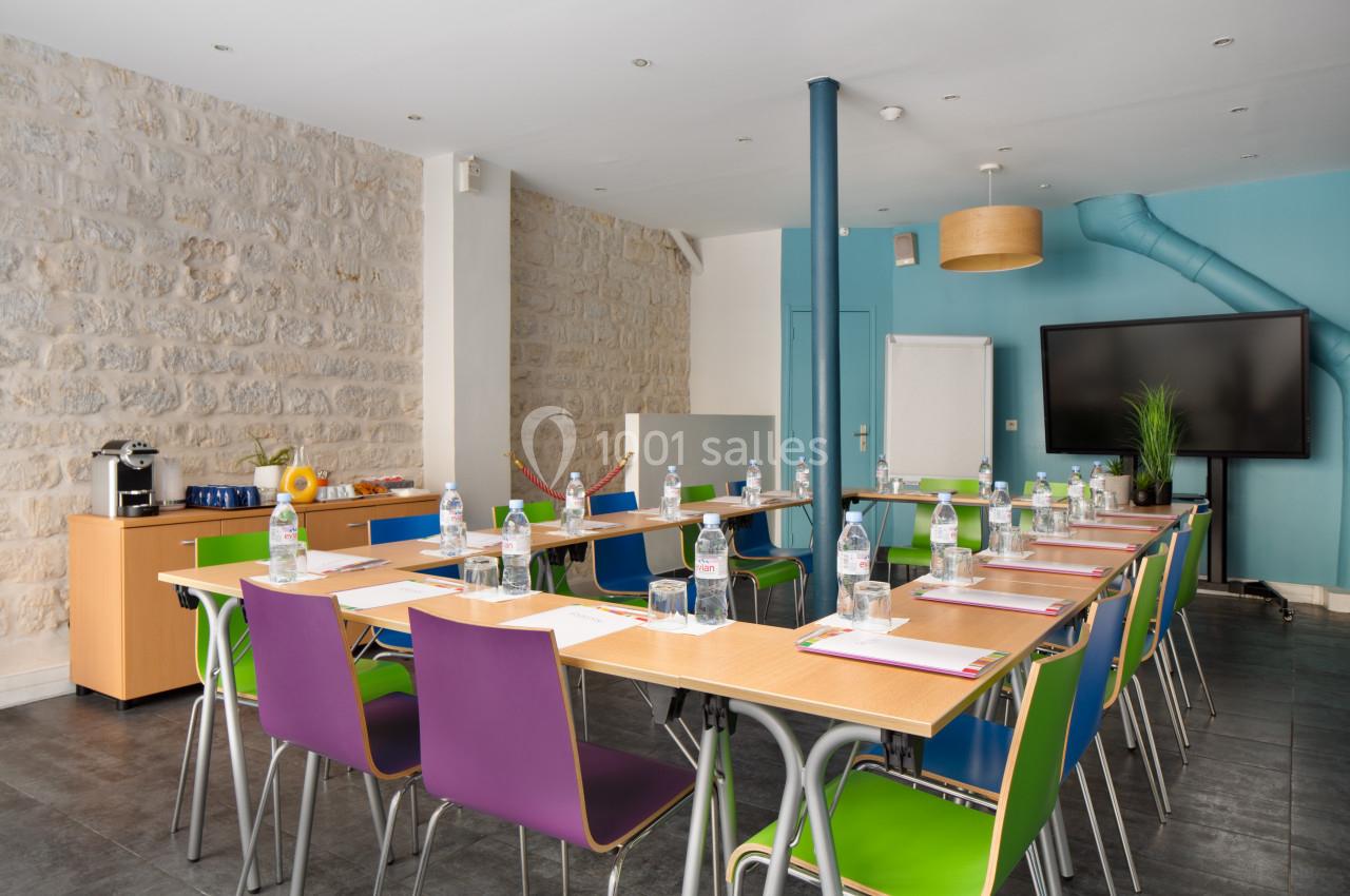 Salle de réunion lumineuse avec tables disposées en U, chaises colorées, écran, et coin rafraîchissements.