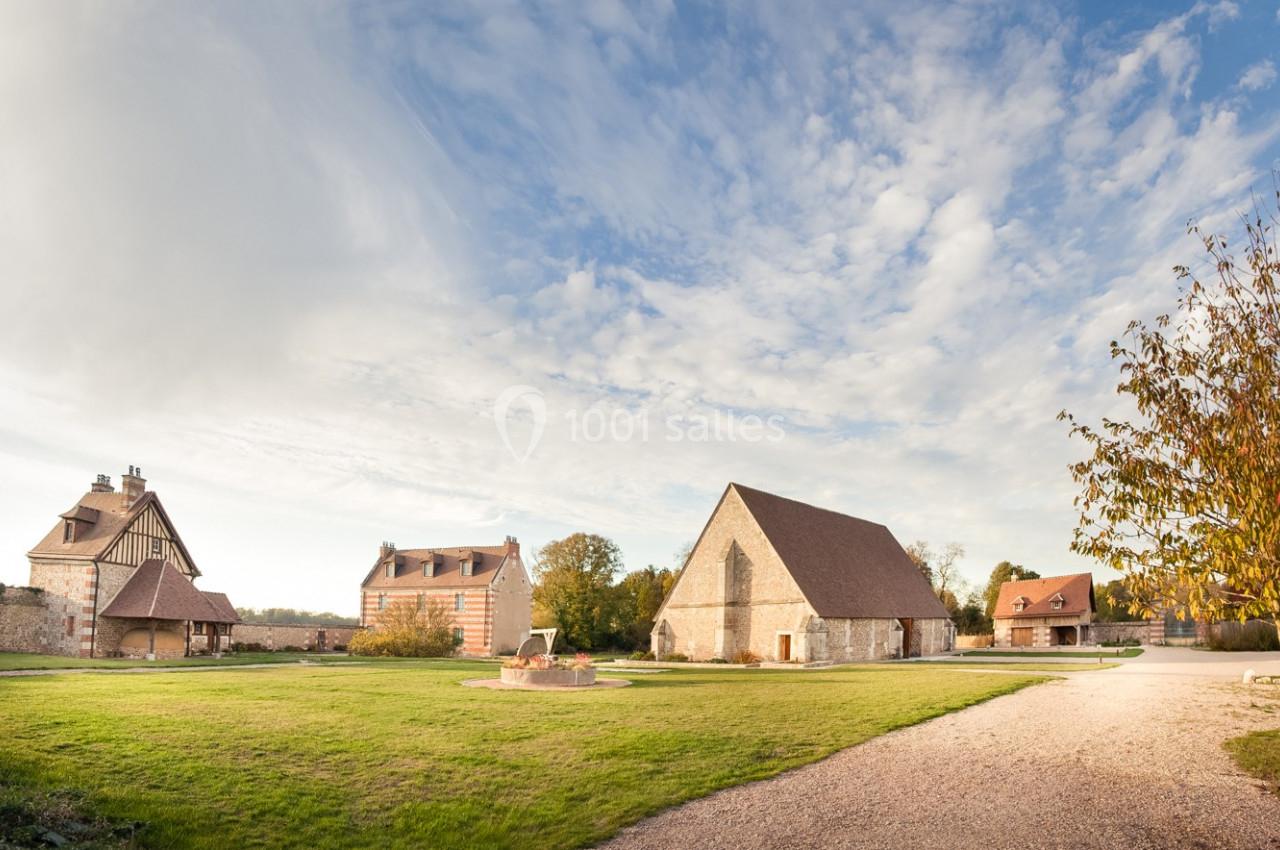 Vue d'un domaine rural avec bâtiments anciens en pierre, pelouse, chemin de gravier et ciel partiellement nuageux.