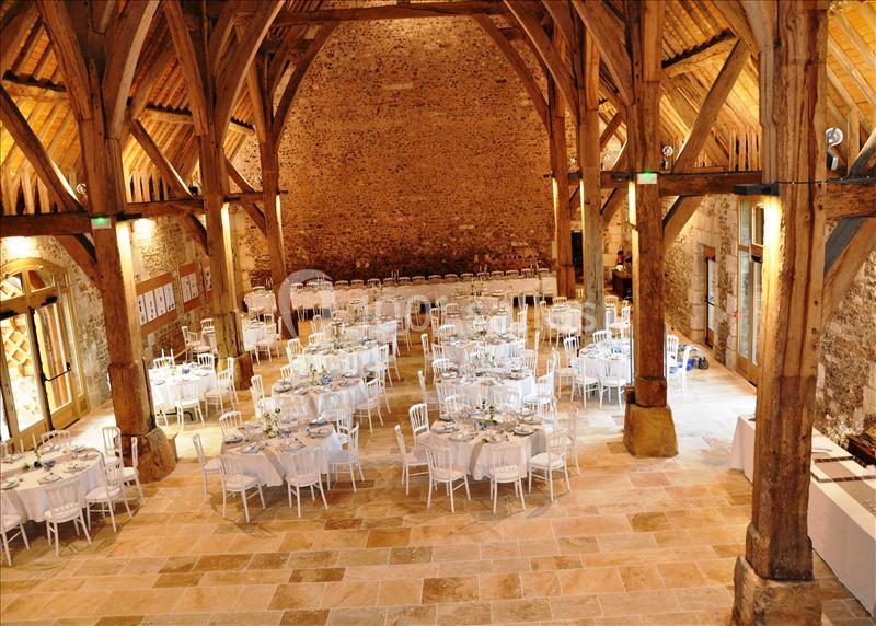 Salle de réception en pierre avec poutres apparentes, tables rondes dressées pour un événement.