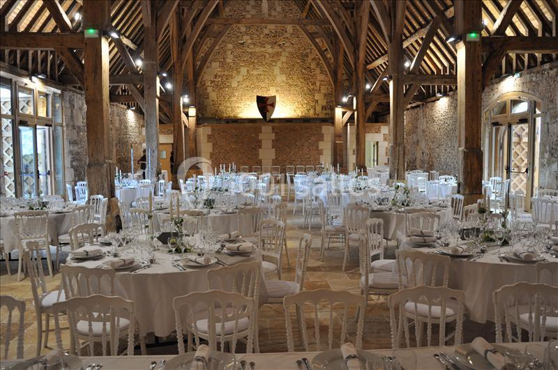 Salle de réception avec tables rondes dressées, chaises blanches et poutres apparentes dans un cadre rustique.