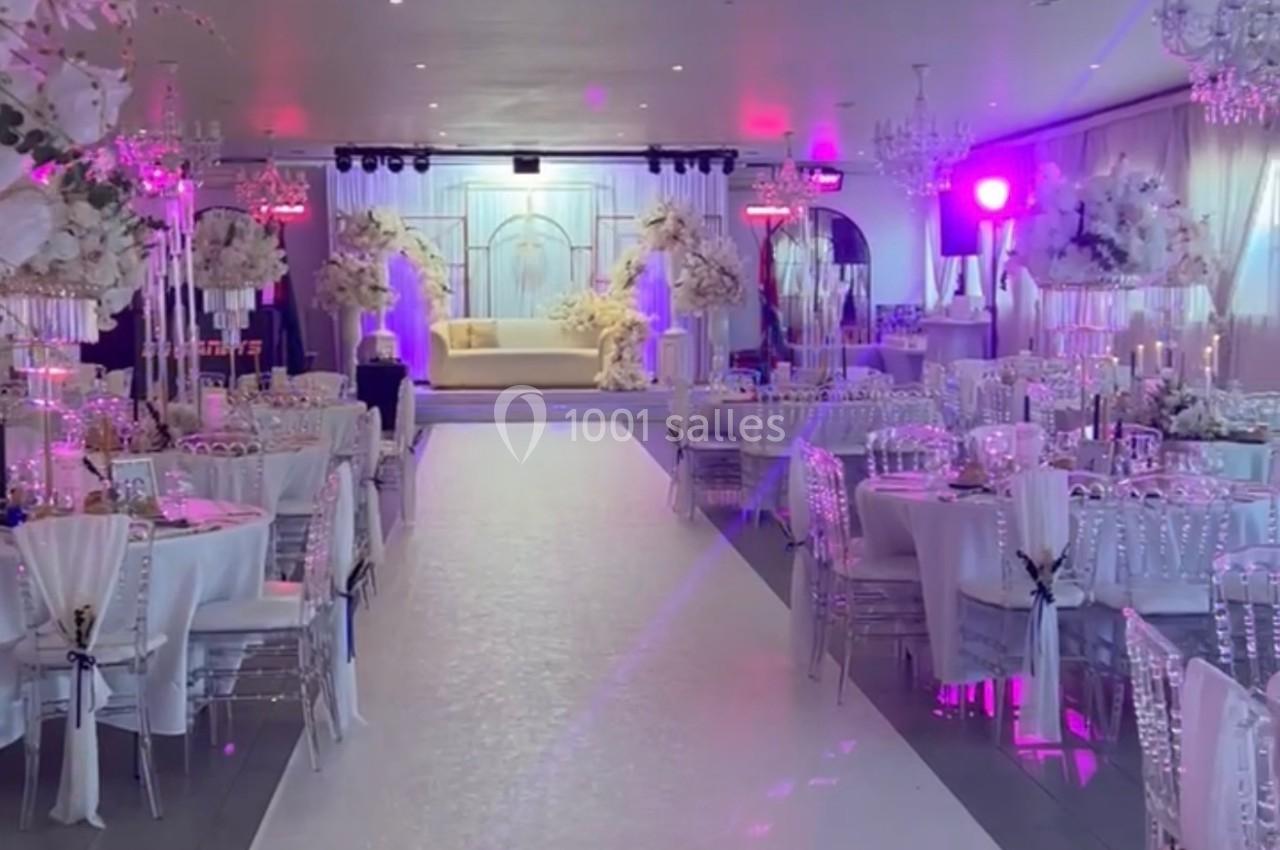 Location salle Beauchamp (Val-d'Oise) - Les Salons Beauchamp #13 Salle de réception décorée avec des fleurs blanches, tables dressées et éclairage rose, allée centrale menant à une scène.