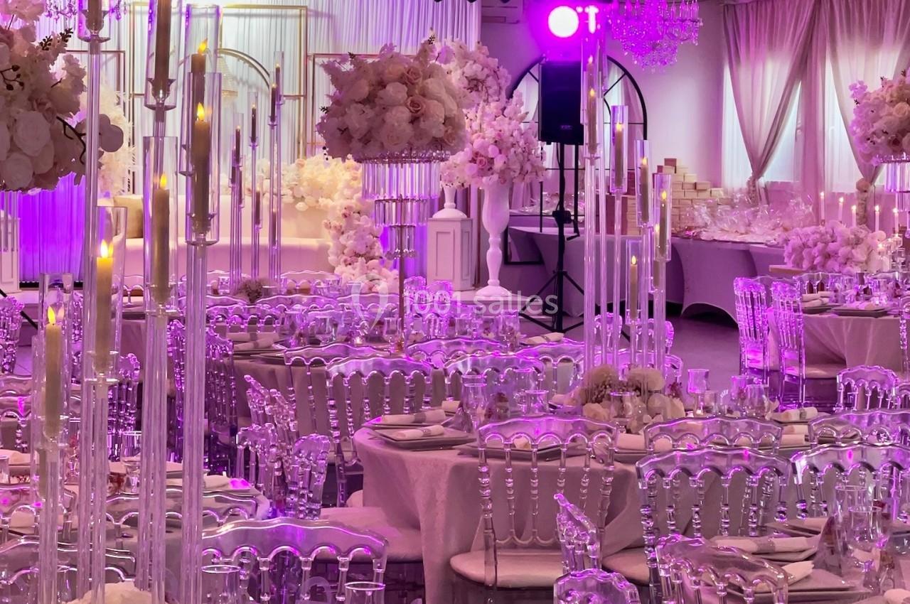 Location salle Beauchamp (Val-d'Oise) - Les Salons Beauchamp #15 Salle de réception décorée avec des chandeliers, des fleurs blanches et des chaises transparentes sous un éclairage rose.