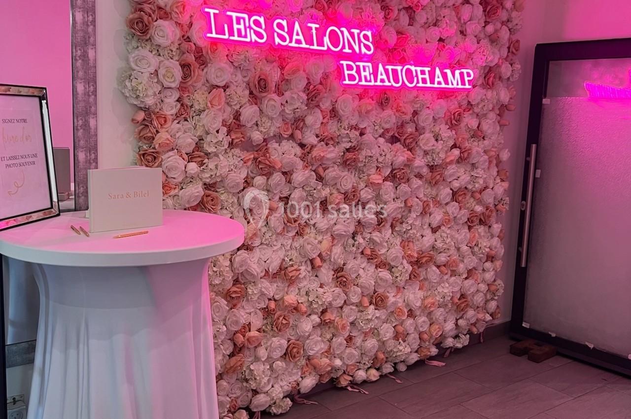 Location salle Beauchamp (Val-d'Oise) - Les Salons Beauchamp #38 Mur de fleurs roses et blanches avec une enseigne néon rose ’Les Salons Beauchamp’, table haute drapée à gauche.