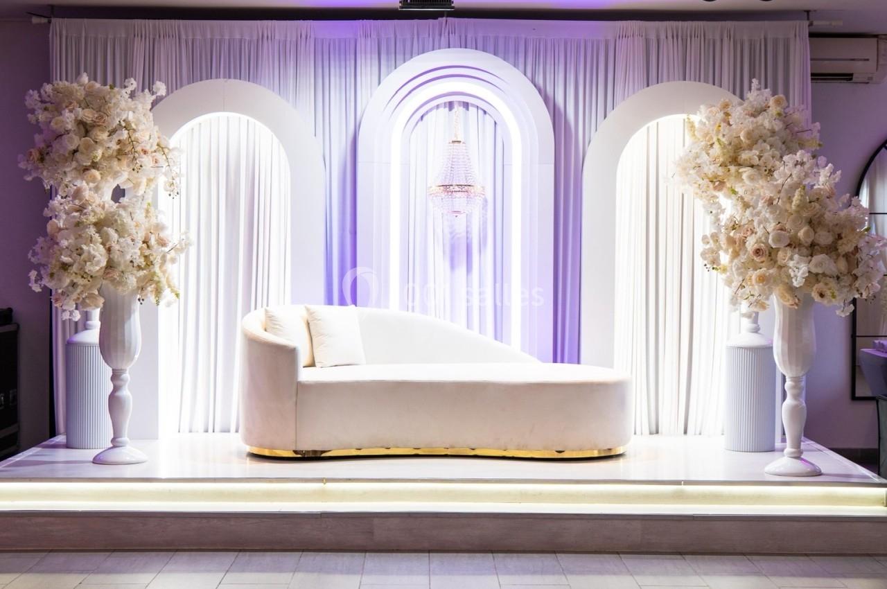 Location salle Beauchamp (Val-d'Oise) - Les Salons Beauchamp #12 Canapé blanc sur une scène décorée de rideaux, arches lumineuses et arrangements floraux blancs.