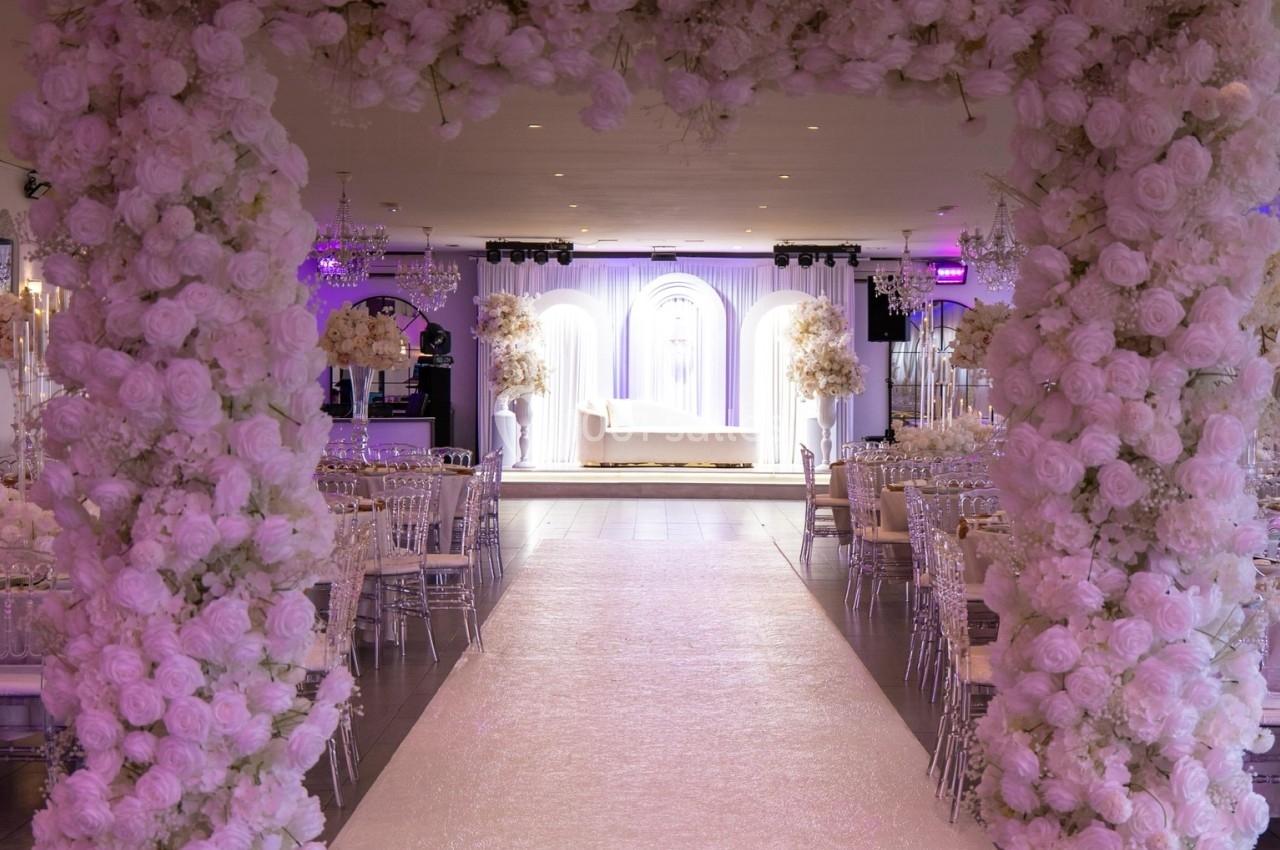 Location salle Beauchamp (Val-d'Oise) - Les Salons Beauchamp #5 Arche florale blanche encadrant une allée menant à une scène décorée pour un événement festif en intérieur.