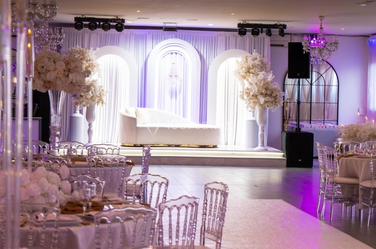 Location salle Beauchamp (Val-d'Oise) - Les Salons Beauchamp #1 Salle de réception élégante avec tables décorées, chaises transparentes et éclairage violet, devant une scène ornée d'arches.
