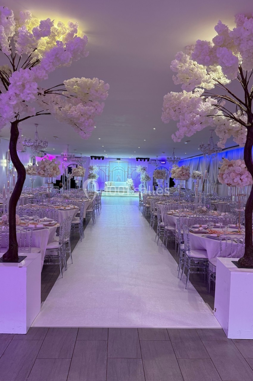 Salle de réception décorée avec des tables dressées, un tapis blanc central et des arbres artificiels fleuris.