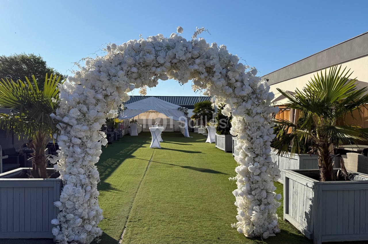 Arche de fleurs blanches sur une pelouse, menant à une tente blanche dans un espace extérieur ensoleillé.