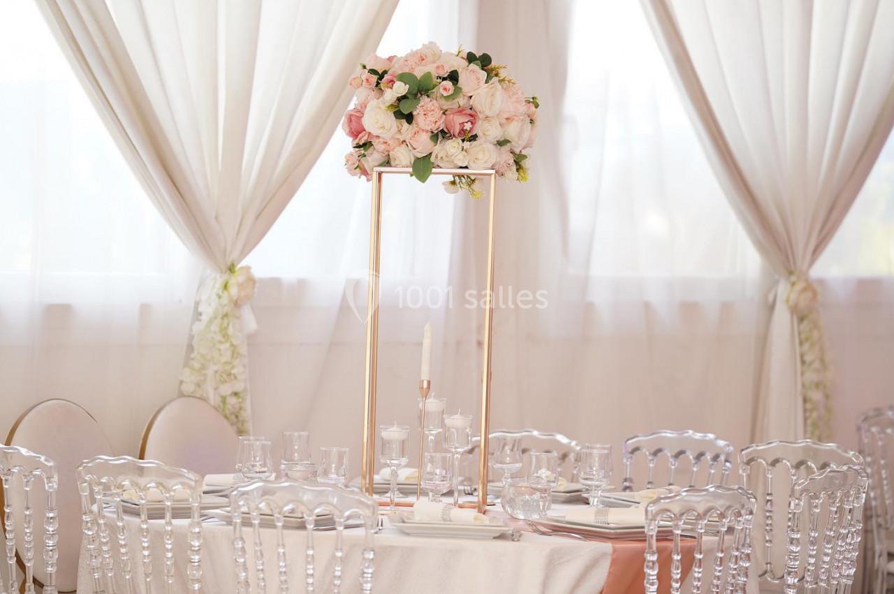 CENTRE DE TABLE Centre de table floral avec roses blanches et roses sur un support doré, entouré de chaises transparentes et nappes blanches.