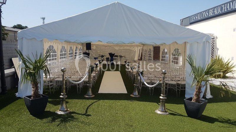 CEREMONIE ESPACE COUVERT Tente blanche aménagée avec des rangées de chaises transparentes, allée centrale beige et décorations végétales sur gazon.