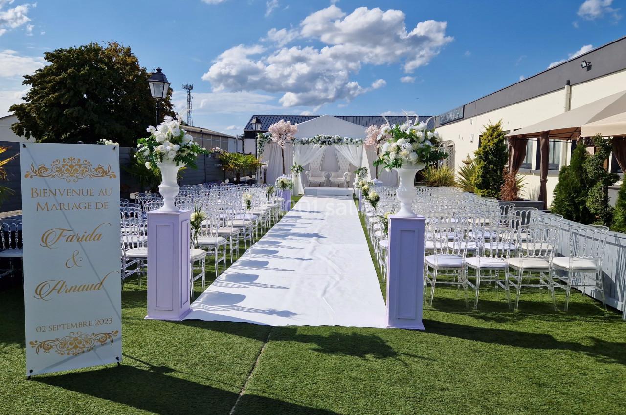 CEREMONIE EXTERIEUR Allée décorée pour une cérémonie de mariage en extérieur, avec chaises transparentes et arche florale au fond.