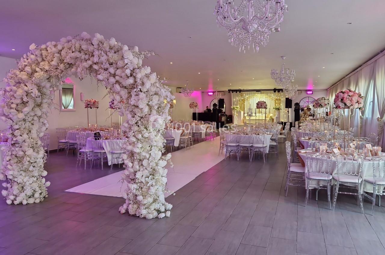 DECO ARCHE FLORALE Salle de réception décorée avec des tables élégantes, un arche floral blanc et un éclairage rose tamisé.
