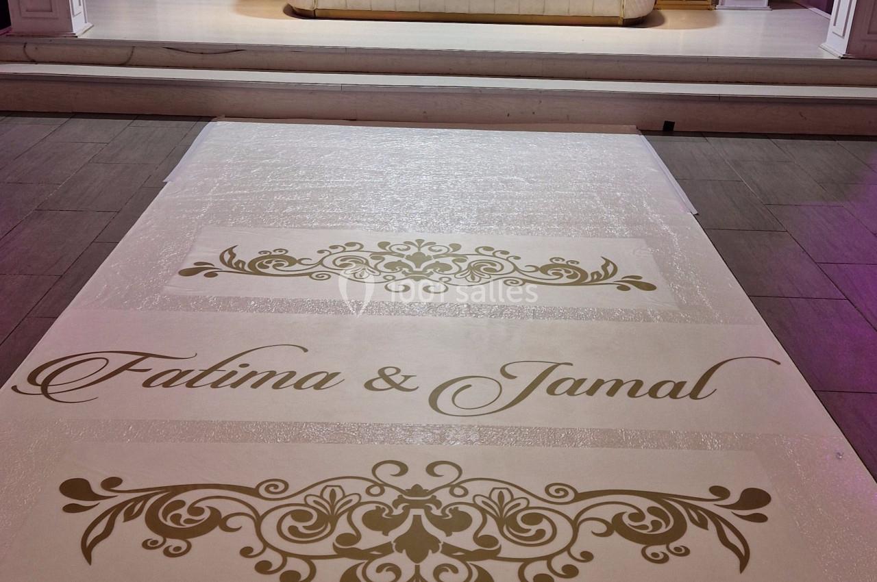 DECORATION PERSONNALISEE Allée décorative avec noms inscrits en doré menant à un canapé blanc devant une arche ornée de fleurs blanches.