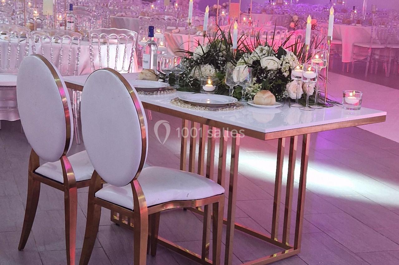 TABLE D HONNEUR COMPOS FLORALE Table décorée avec des bougies et des fleurs, entourée de chaises blanches, dans une salle de réception élégante.