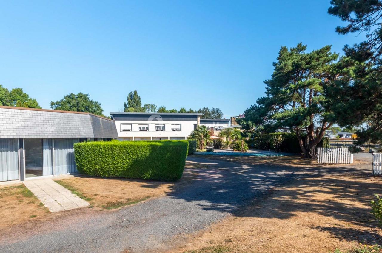 Location salle Saint-Aignan-de-Cramesnil (Calvados) - La Jalousie #8 Vue d'un bâtiment moderne entouré d'arbres et de haies, avec une allée en gravier et une pelouse partiellement sèche.