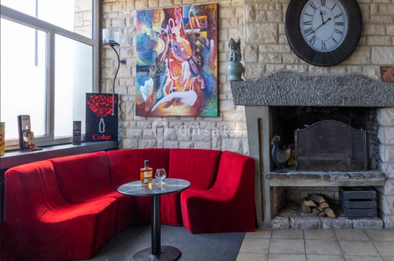Location salle Saint-Aignan-de-Cramesnil (Calvados) - La Jalousie #4 Canapé rouge en angle autour d'une table basse, cheminée en pierre et horloge murale dans un salon lumineux.