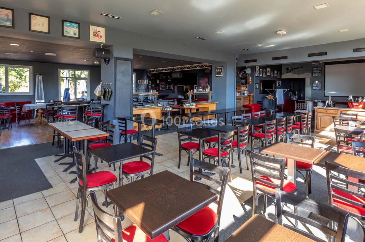 Location salle Saint-Aignan-de-Cramesnil (Calvados) - La Jalousie #2 Salle de restaurant lumineuse avec des tables et chaises noires, assises rouges, et une scène en arrière-plan.