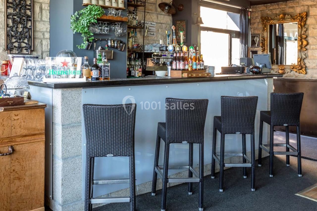 Location salle Saint-Aignan-de-Cramesnil (Calvados) - La Jalousie #6 Comptoir de bar moderne avec tabourets en osier, étagères garnies de bouteilles et décoration murale en pierre.