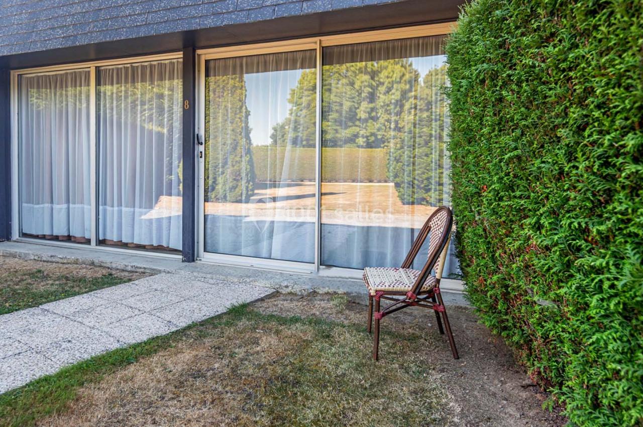 Location salle Saint-Aignan-de-Cramesnil (Calvados) - La Jalousie #19 Chaise en osier placée près d'une haie verte devant une maison avec de grandes baies vitrées et un chemin pavé.