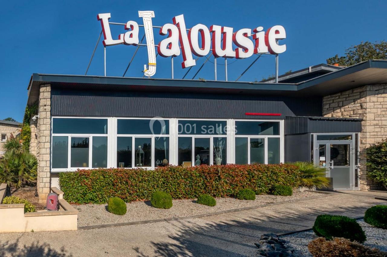 Location salle Saint-Aignan-de-Cramesnil (Calvados) - La Jalousie #7 Façade d'un bâtiment avec une enseigne ’La Jalousie’, entourée de plantes et d'arbustes sous un ciel bleu.