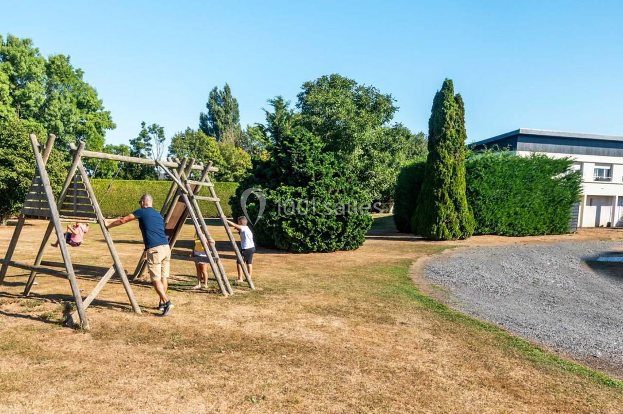Location salle Saint-Aignan-de-Cramesnil (Calvados) - La Jalousie #11 Un adulte et deux enfants jouent sur une balançoire en bois dans un espace extérieur avec arbres et pelouse.