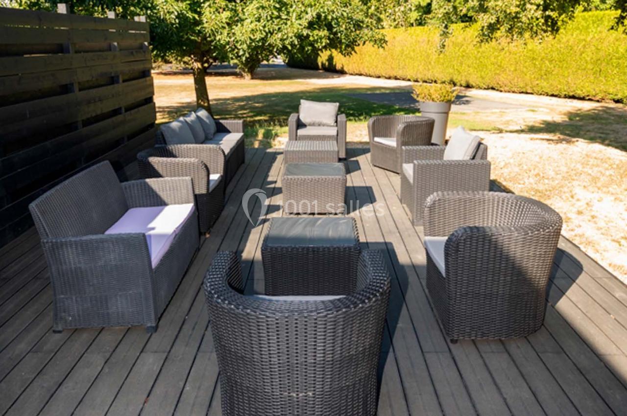 Location salle Saint-Aignan-de-Cramesnil (Calvados) - La Jalousie #16 Salon de jardin en résine tressée avec fauteuils et canapés disposés en cercle sur une terrasse en bois, entourée de verdure.