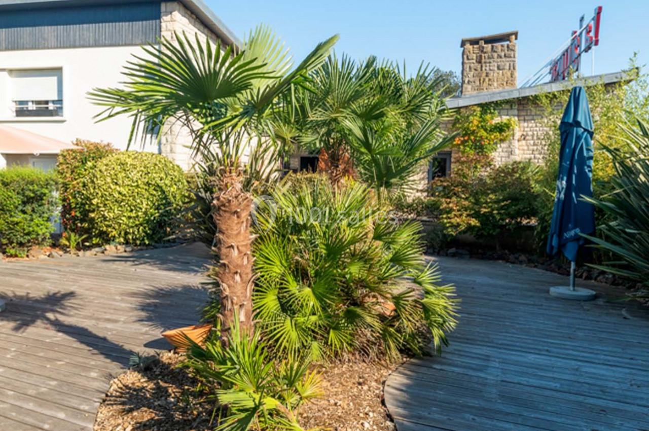 Location salle Saint-Aignan-de-Cramesnil (Calvados) - La Jalousie #17 Terrasse en bois entourée de palmiers et de végétation, avec un parasol bleu et un bâtiment en arrière-plan.