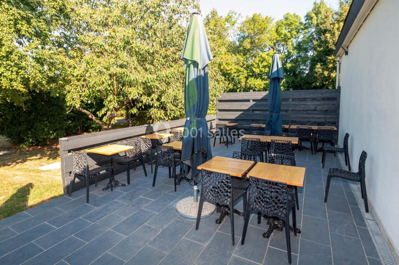Location salle Saint-Aignan-de-Cramesnil (Calvados) - La Jalousie #15 Terrasse extérieure avec tables en bois, chaises noires et parasols, entourée de verdure et d'une clôture en bois.