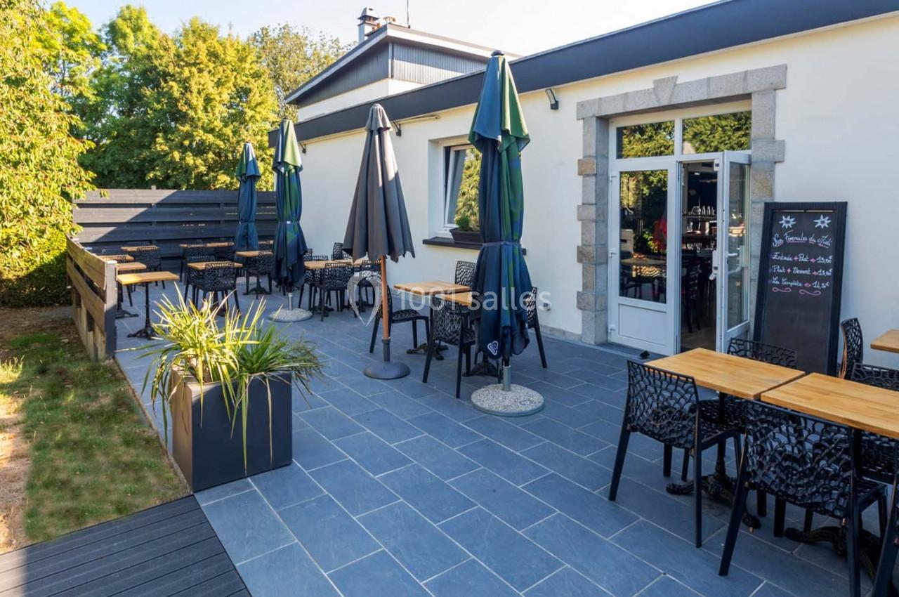 Location salle Saint-Aignan-de-Cramesnil (Calvados) - La Jalousie #10 Terrasse extérieure d'un restaurant avec tables, chaises et parasols, entourée de verdure et d'un bâtiment clair.
