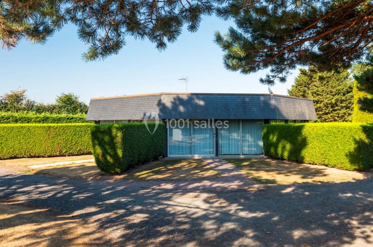 Location salle Saint-Aignan-de-Cramesnil (Calvados) - La Jalousie #18 Maison basse avec toit en ardoise, entourée de haies taillées, sous des arbres projetant des ombres au sol.