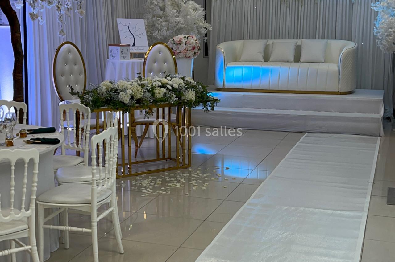 Salle de réception décorée avec des chaises blanches, une table ornée de fleurs et une estrade avec canapé lumineux.