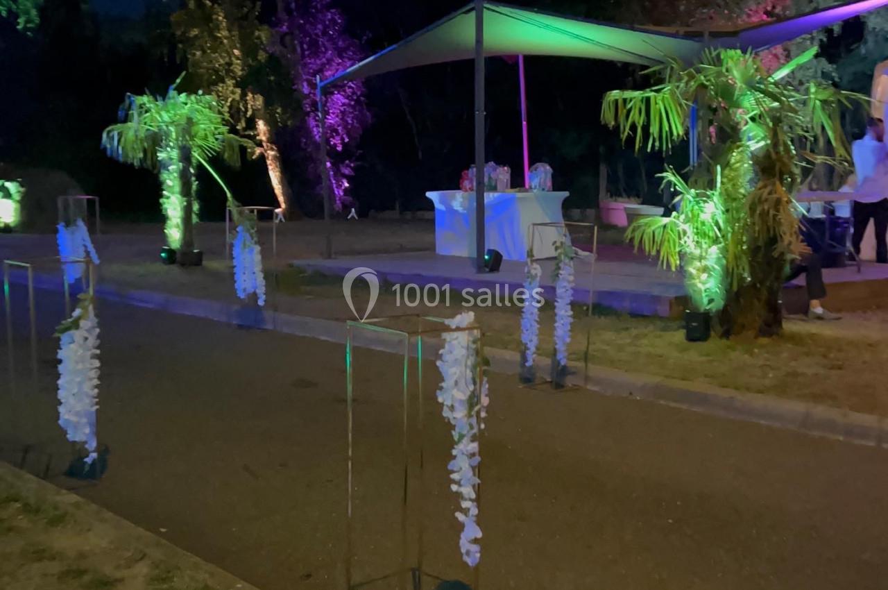Allée décorée de guirlandes lumineuses et végétation tropicale, menant à un espace couvert éclairé de nuit.