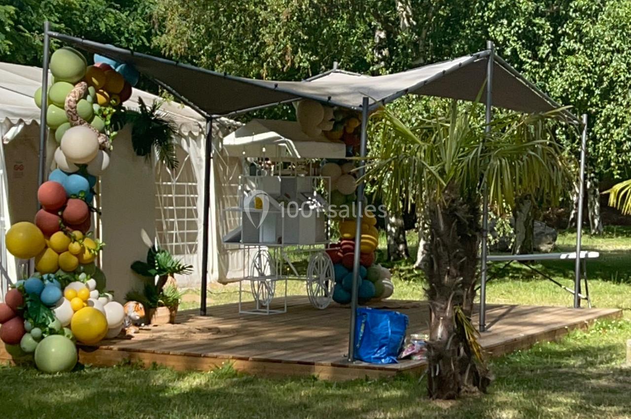 Tente extérieure avec décoration de ballons colorés, mobilier blanc et arbres en arrière-plan dans un espace vert.