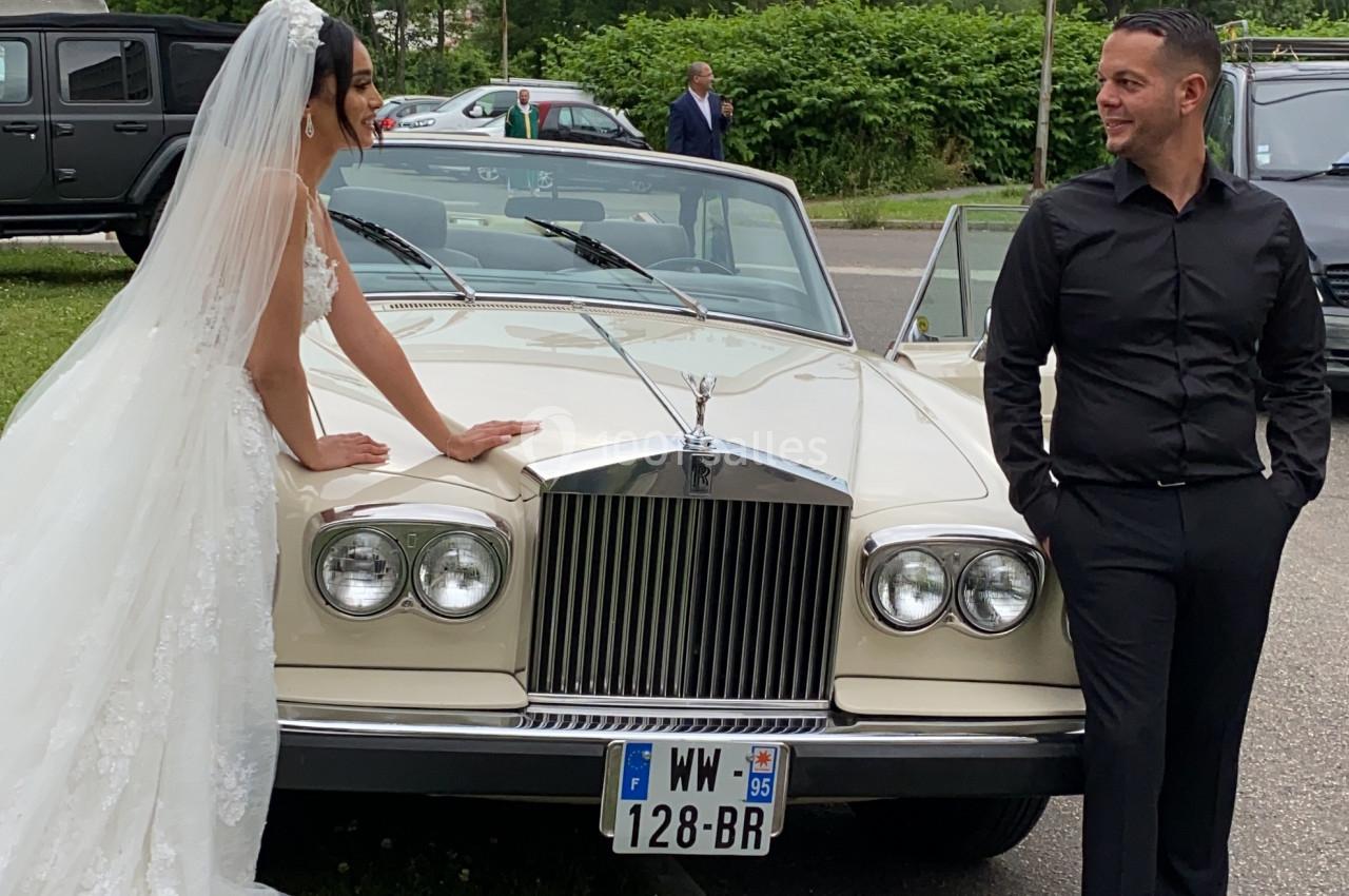Une mariée en robe blanche et un homme en noir posent près d'une voiture ancienne beige, en extérieur.