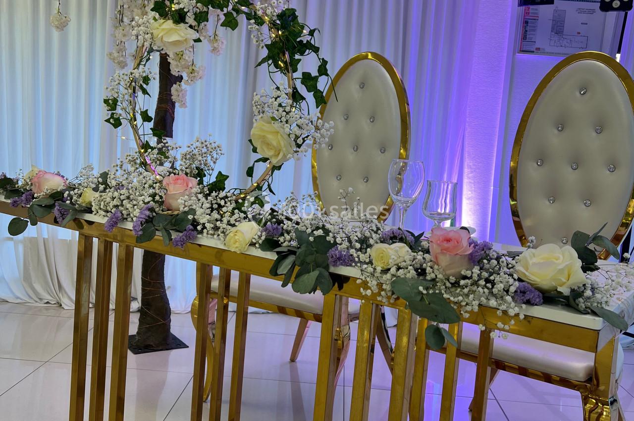 Table décorée avec des fleurs blanches et roses, chaises élégantes et fond lumineux avec rideaux et arbre fleuri.