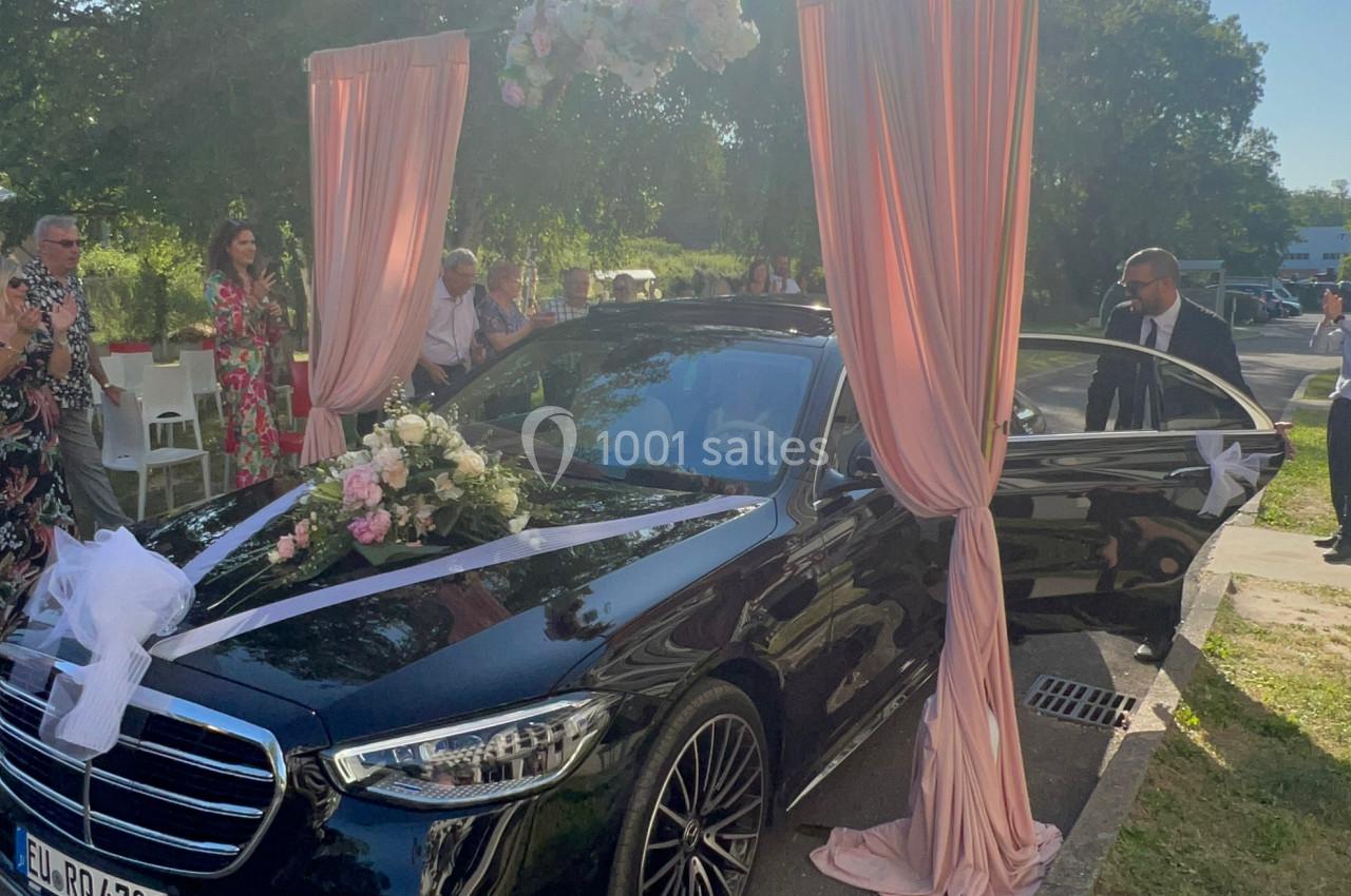 Voiture noire décorée de fleurs et de rideaux roses, garée sur une route lors d'une cérémonie en plein air.