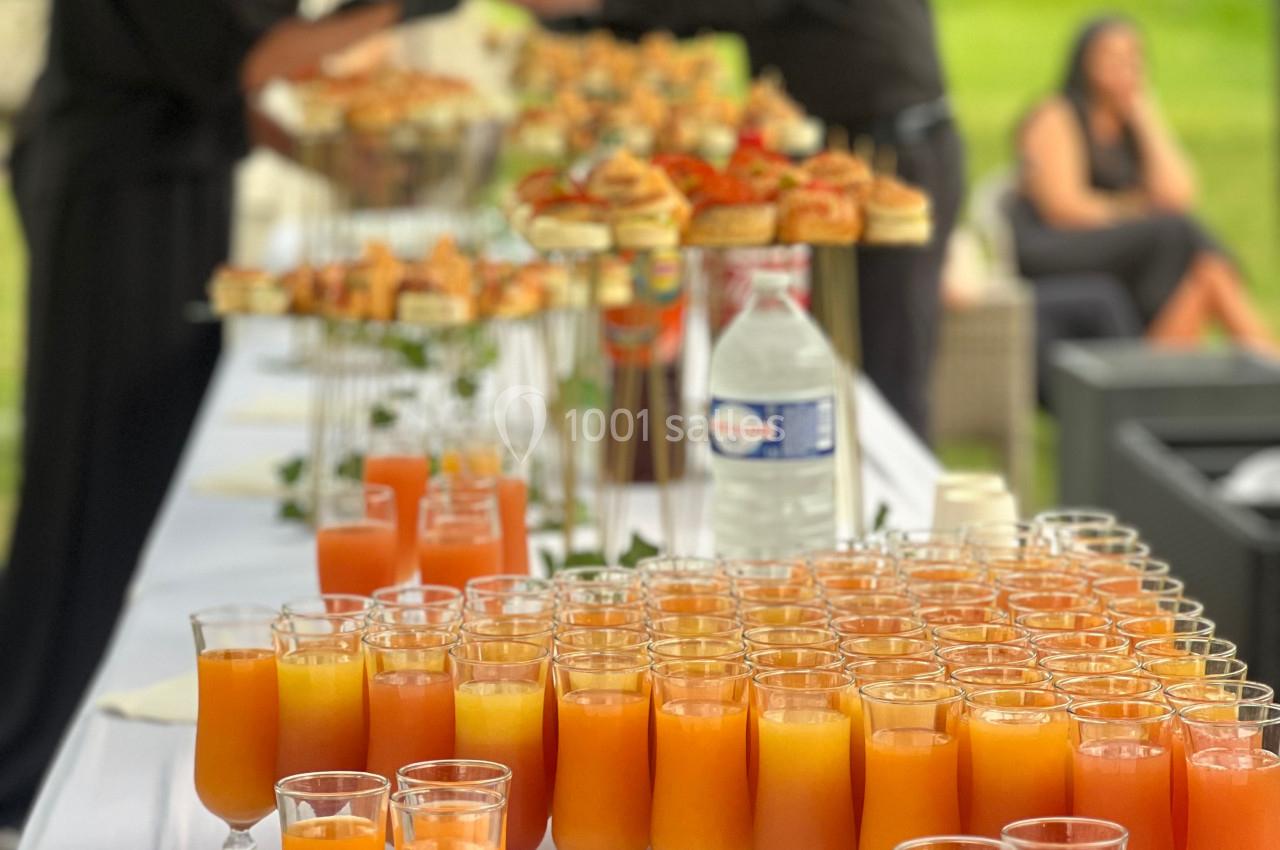Buffet avec verres de jus orange en premier plan et serveurs en arrière-plan dans un cadre extérieur verdoyant.