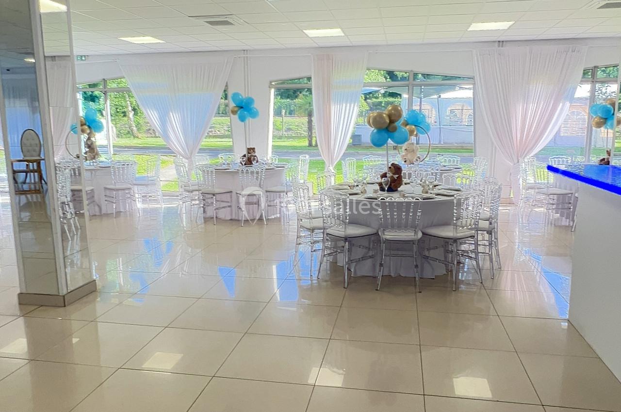 Salle lumineuse avec tables rondes décorées de ballons bleus et blancs, grandes baies vitrées donnant sur un espace vert.