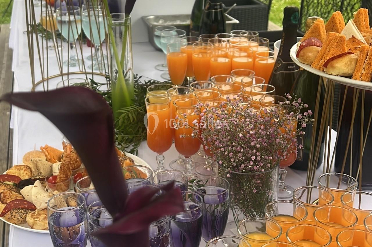 Buffet en extérieur avec verrines, jus colorés, fleurs décoratives et pâtisseries sur une table blanche.