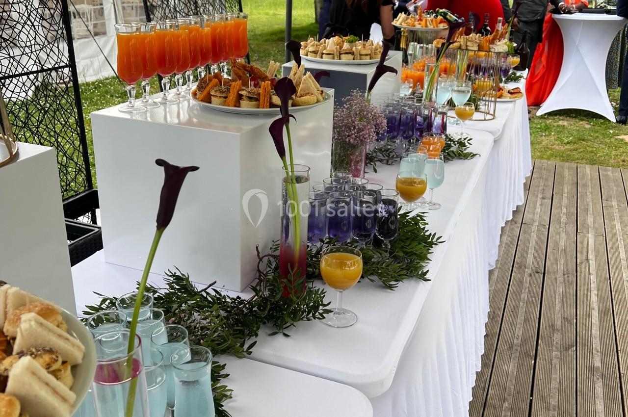 Buffet extérieur avec boissons colorées, verrines et amuse-bouches disposés sur des tables blanches décorées de fleurs.
