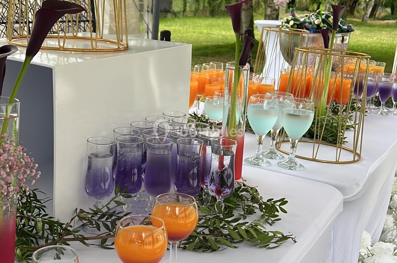Table décorée en extérieur avec des verres colorés, des fleurs et des éléments décoratifs dorés sur une nappe blanche.