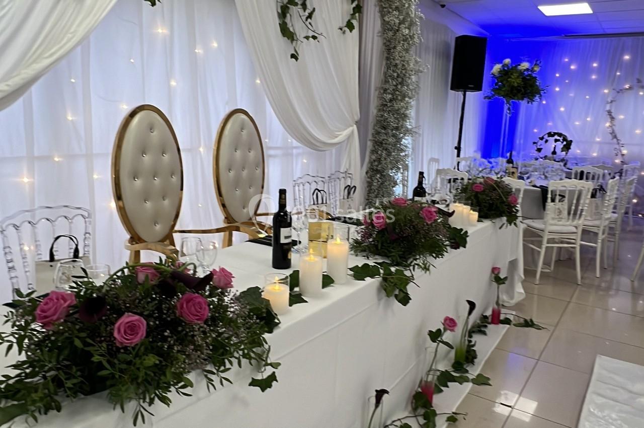 Table décorée pour un événement avec fleurs roses, bougies, chaises blanches et rideaux ornés de guirlandes lumineuses.