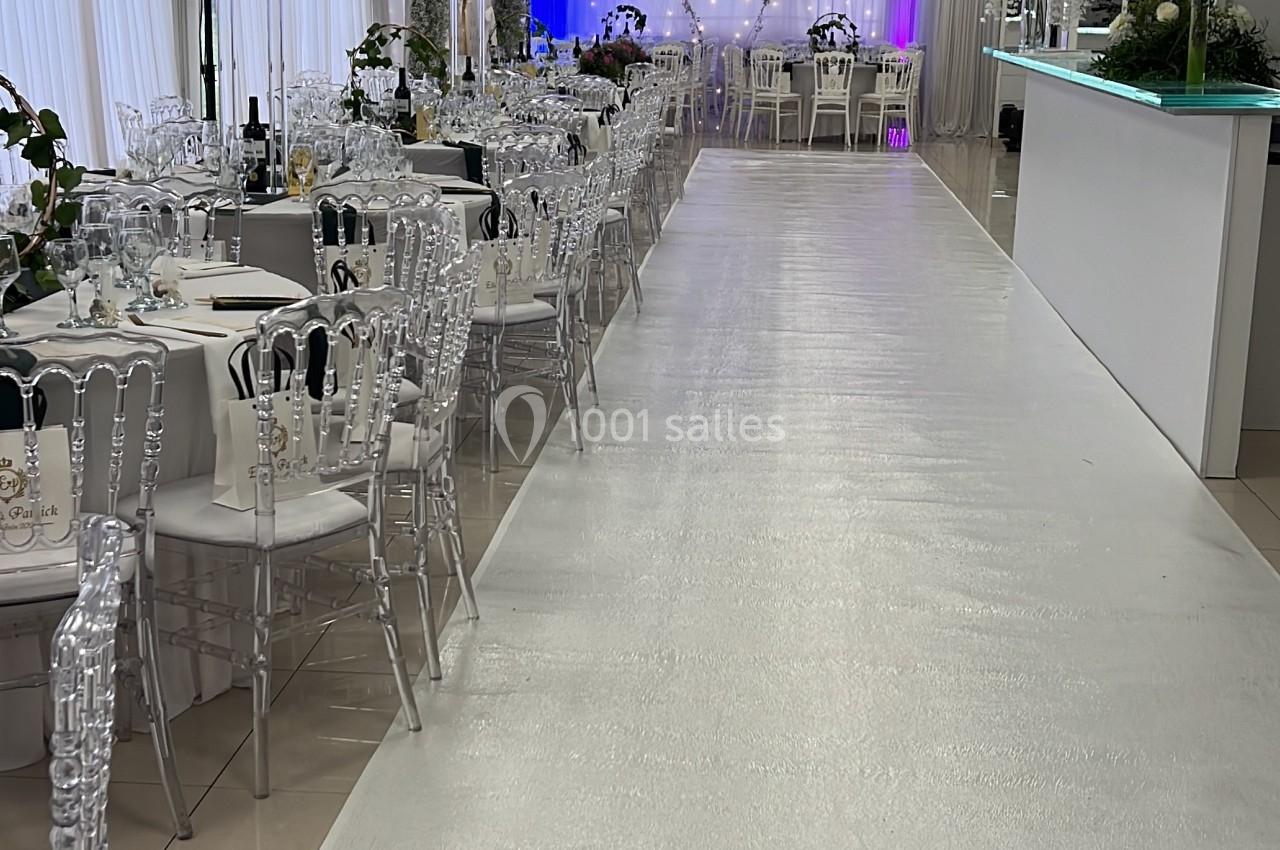 Salle de réception lumineuse avec tables décorées, chaises transparentes et éclairage violet en fond.
